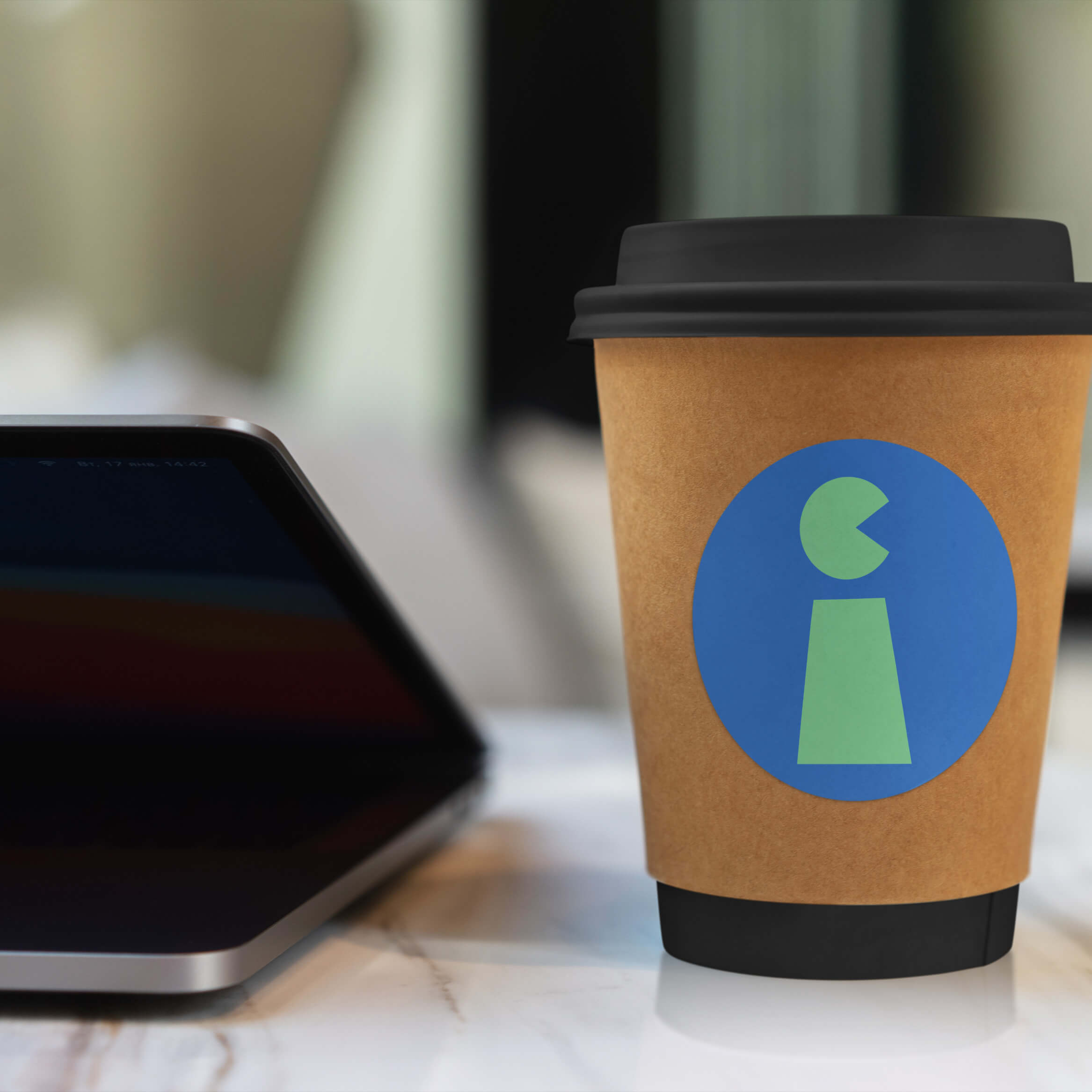 A brown coffee cup with a blue and green geometric sticker next to a laptop.