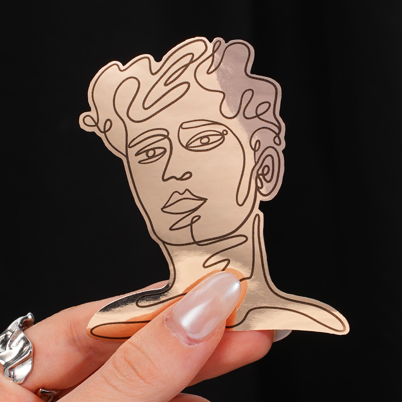 Vinyl sticker with a metallic gold finish and a minimalist line drawing of a face, held between fingers.