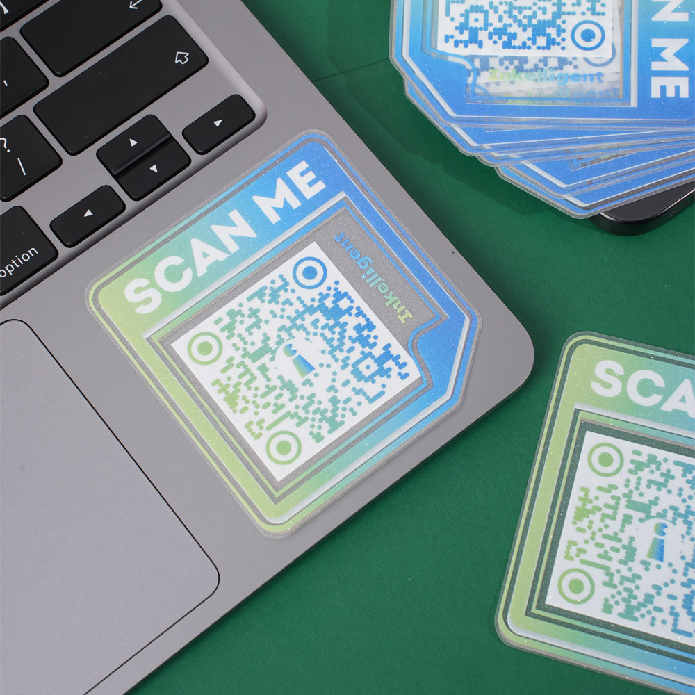 QR code sticker with 'Scan Me' text in gradient blue and green, placed on a laptop near a stack of similar stickers on a green surface.