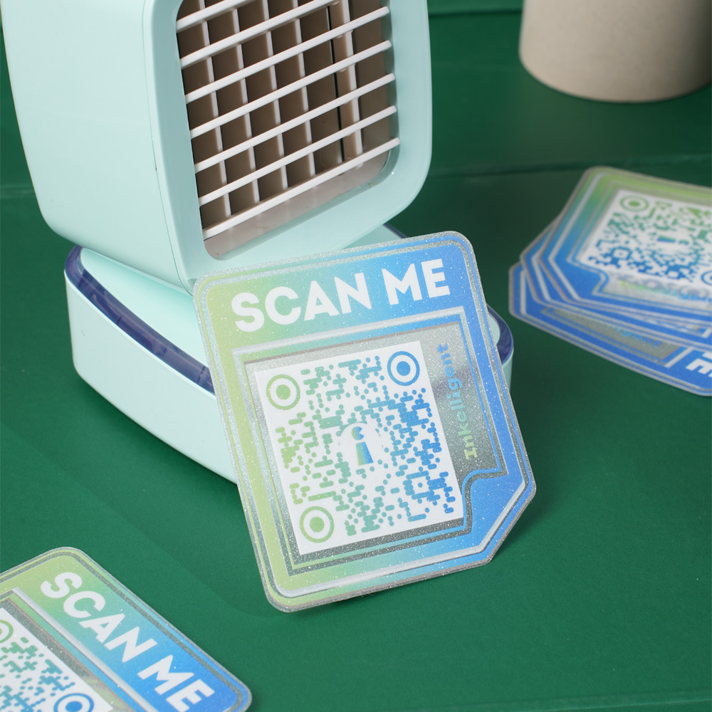 Sticker with a QR code and 'Scan Me' text in gradient blue and green, placed near a small portable fan on a green surface.
